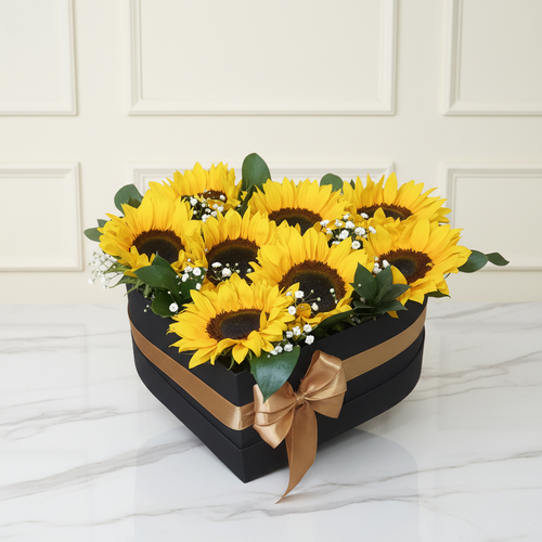 Caja negra en forma de corazón con lazo dorado, llena de girasoles amarillos y flores blancas sobre mesa de mármol