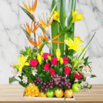 Arreglo de flores amarillas, naranjas y rojas combinado con frutas tropicales como uvas, manzanas y naranjas sobre una base de madera, con fondo de pared de mármol claro