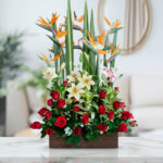 Arreglo floral rectangular con rosas rojas, lirios blancos y rosados y flores aves del paraíso en un jarrón de madera sobre una mesa de mármol