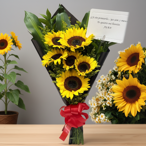 Ramo de girasoles amarillos envueltos en papel negro con lazo rojo y tarjeta de dedicatoria sobre mesa de madera, con macetas de girasoles de fondo