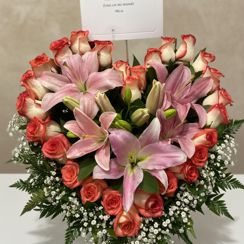 Arreglo floral en forma de corazón con lirios rosados y rosas rojas y blancas, decorado con gypsophila y follaje verde, con tarjeta en la parte superior