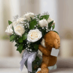 Ramo de rosas blancas y flores pequeñas en jarrón de cristal con lazo blanco acompañado de oso de peluche marrón
