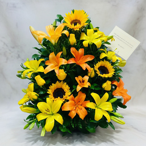 Arreglo floral en forma de pirámide con lirios amarillos y naranjas y girasoles sobre fondo claro