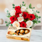 Arreglo floral de rosas rojas y blancas con follaje verde acompañado de una caja de chocolates Ferrero Rocher sobre superficie de mármol