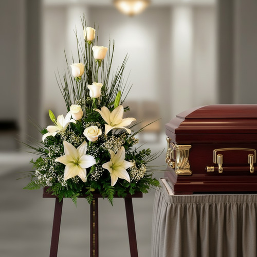 Arreglo floral fúnebre con lirios y rosas blancas junto a un ataúd de madera barnizada en una sala velatoria