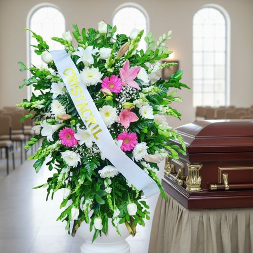 Arreglo floral funerario grande con flores blancas y rosas y cinta con inscripción, colocado junto a un ataúd de madera en una capilla luminosa