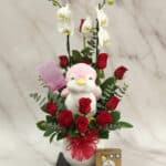 Arreglo floral ALICE con orquídeas blancas, rosas rojas frescas, peluche de pingüino rosa y chocolates Ferrero Rocher, ideal