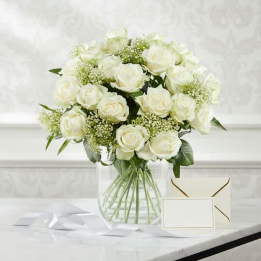 Arreglo floral Estefanía con 24 rosas blancas frescas en jarrón de cristal, tarjeta de regalo y cinta satinada sobre mármol.