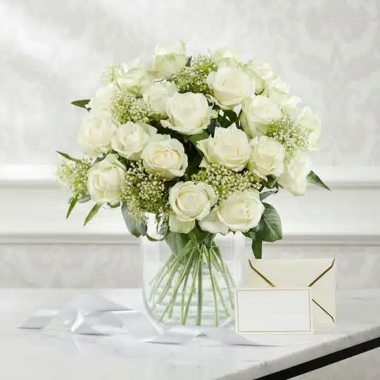 Arreglo floral Estefanía con 24 rosas blancas frescas en jarrón de cristal, tarjeta de regalo y cinta satinada sobre mármol.
