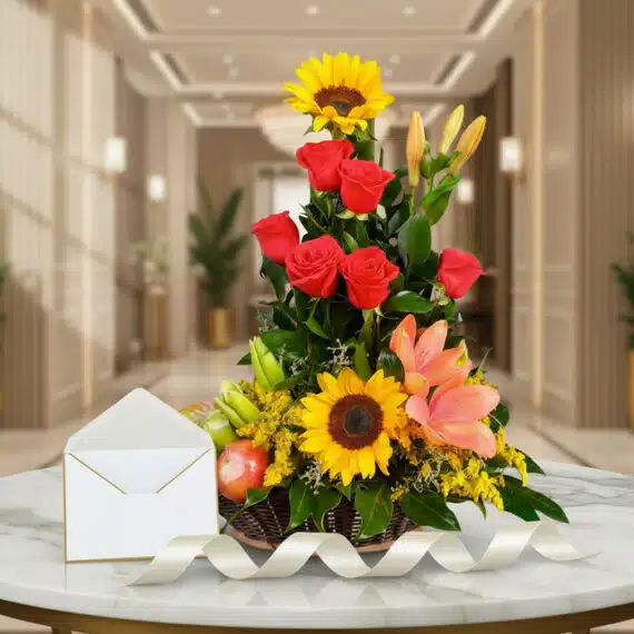 Arreglo floral con rosas rojas, girasoles, lirios y frutas kiwano sobre una mesa de mármol, con una cinta decorativa.