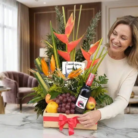 Mujer sonriente admirando un arreglo floral con heliconias, frutas frescas y una botella de vino tinto, presentado en una caj