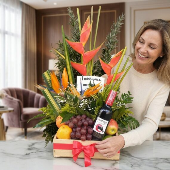 Mujer sonriente admirando un arreglo floral con heliconias, frutas frescas y una botella de vino tinto, presentado en una caj
