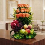 Arreglo floral premium con rosas salmón, uvas, manzanas, duraznos y peras, sobre una mesa de madera pulida en un lobby de hot