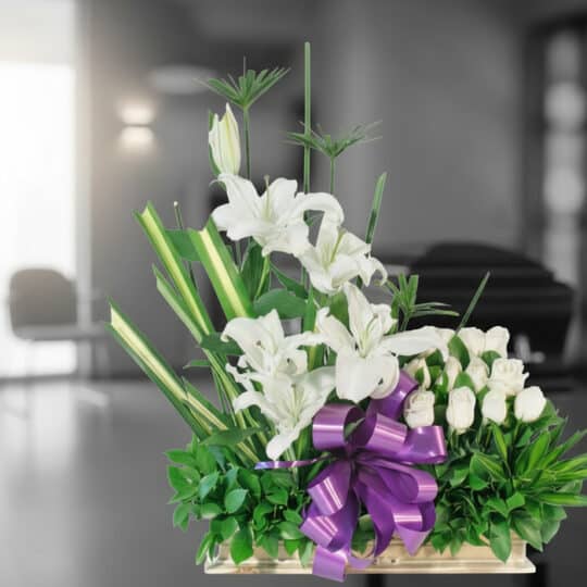 Arreglo fúnebre Pureza con lirios y rosas blancas, lazo morado, en sala de velación moderna con ataúd desenfocado al fondo.