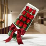 Caja de 12 rosas rojas frescas y vibrantes, presentadas en un elegante estuche blanco con un lazo de satén rojo, sobre una su