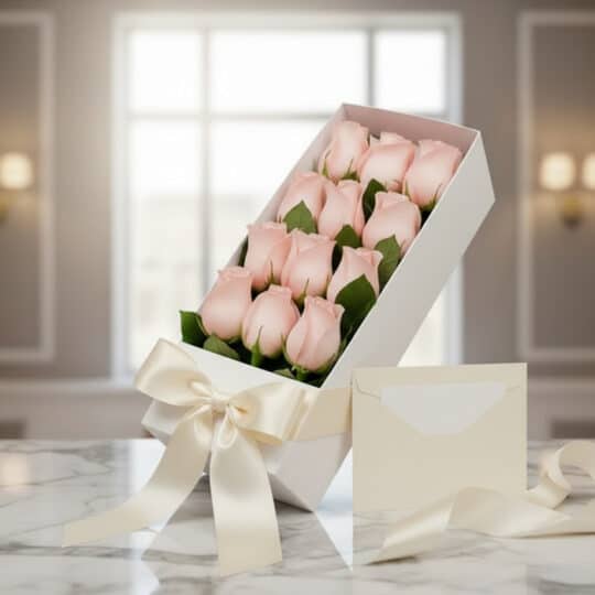 Caja blanca inclinada con doce rosas rosadas frescas, lazo de satén color crema y tarjeta de regalo sobre mármol pulido, con