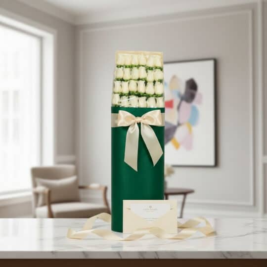 Caja cilíndrica verde con 24 rosas blancas, lazo satinado color crema, tarjeta de regalo y cinta sobre mármol.