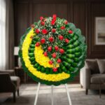 Corona fúnebre Adela con rosas rojas y flores blancas sobre base de crisantemos amarillos y hojas verdes, en sala de velación