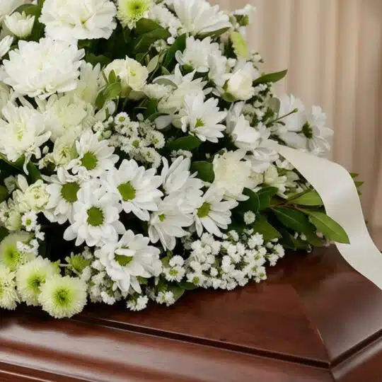 Primer plano de un arreglo floral fúnebre blanco y verde, con rosas, crisantemos y follaje, sobre una caja de madera oscura,