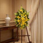 Pedestal de homenaje "El Legado de Sebastian" con un arreglo floral fúnebre de lirios y crisantemos amarillos, presentado ele